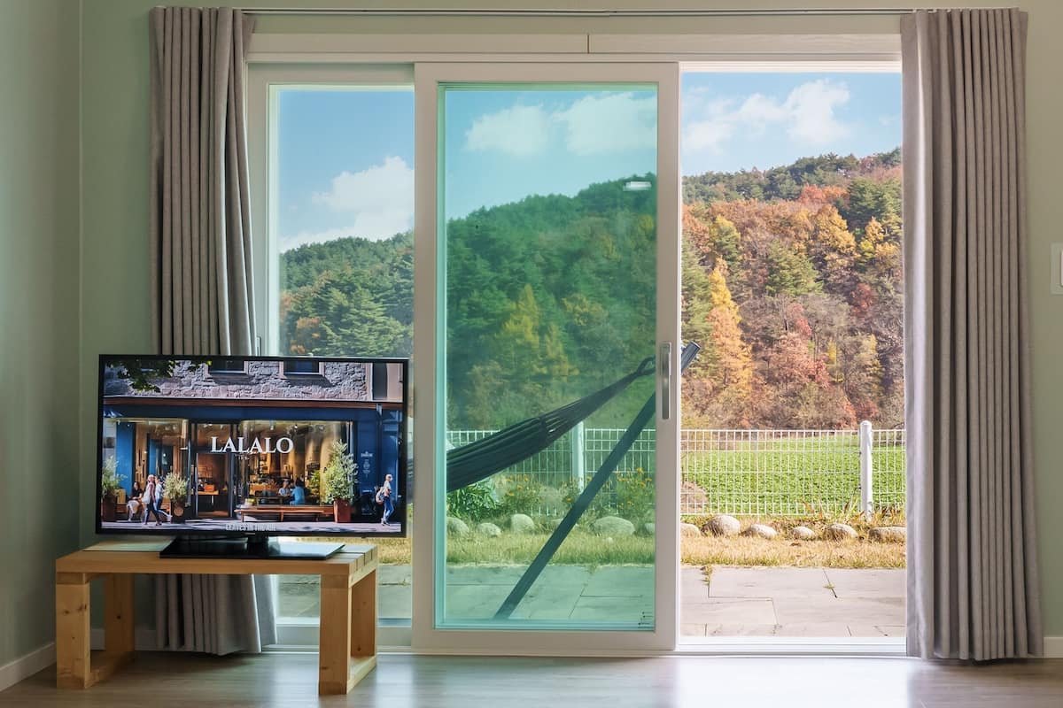 Una habitació amb televisió que mostra una botiga anomenada "LAJALO", una porta corredissa de vidre que dona a un jardí amb cadira espanyola, i vista a un turó boscós.