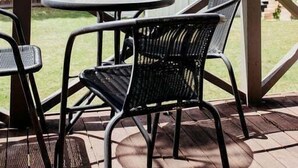 An outdoor patio with a round table and two chairs, a red polka-dotted bowl with flowers, and a view of a house and trees. - StudioNESS Scamander (Break O'Day)