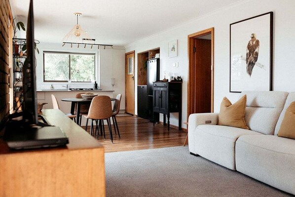 A modern living room with a sofa, a dining area with chairs, a kitchen with cabinets, and a large window.