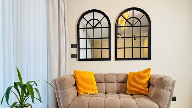 A beige sofa with two yellow cushions, a potted plant, and a glass table with books. Two black-framed windows with a grid pattern are on the wall. - El mejor departamento de Macul (Santiago)