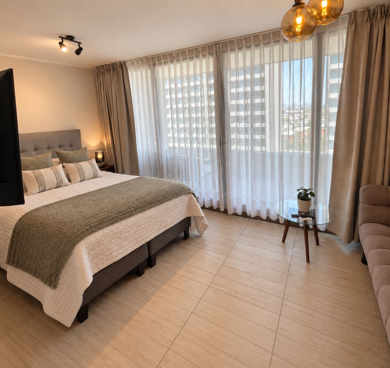 A modern hotel room with a large bed, a round glass table, and a view of the city through sheer curtains.
