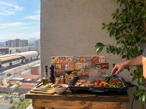 Une personne qui fait griller de la nourriture sur un barbecue extérieur, avec une vue sur la ville en arrière-plan.