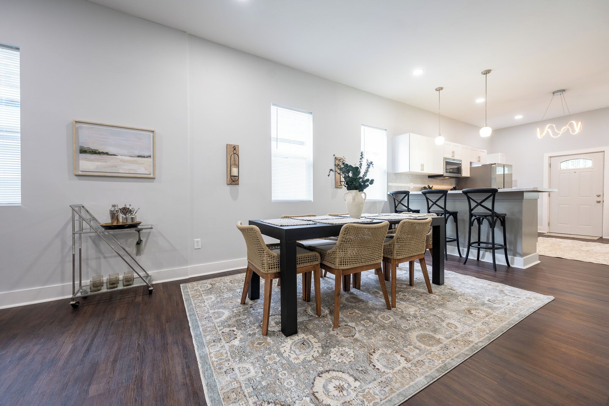 A modern kitchen with white cabinets, a dining area with a black table and wicker chairs, and a patterned rug on the floor.