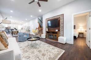 A spacious living room with a large TV, comfortable seating, and a staircase leading to another level. -  Spacious 5-Bedroom Blue Bungalow (Indianapolis)