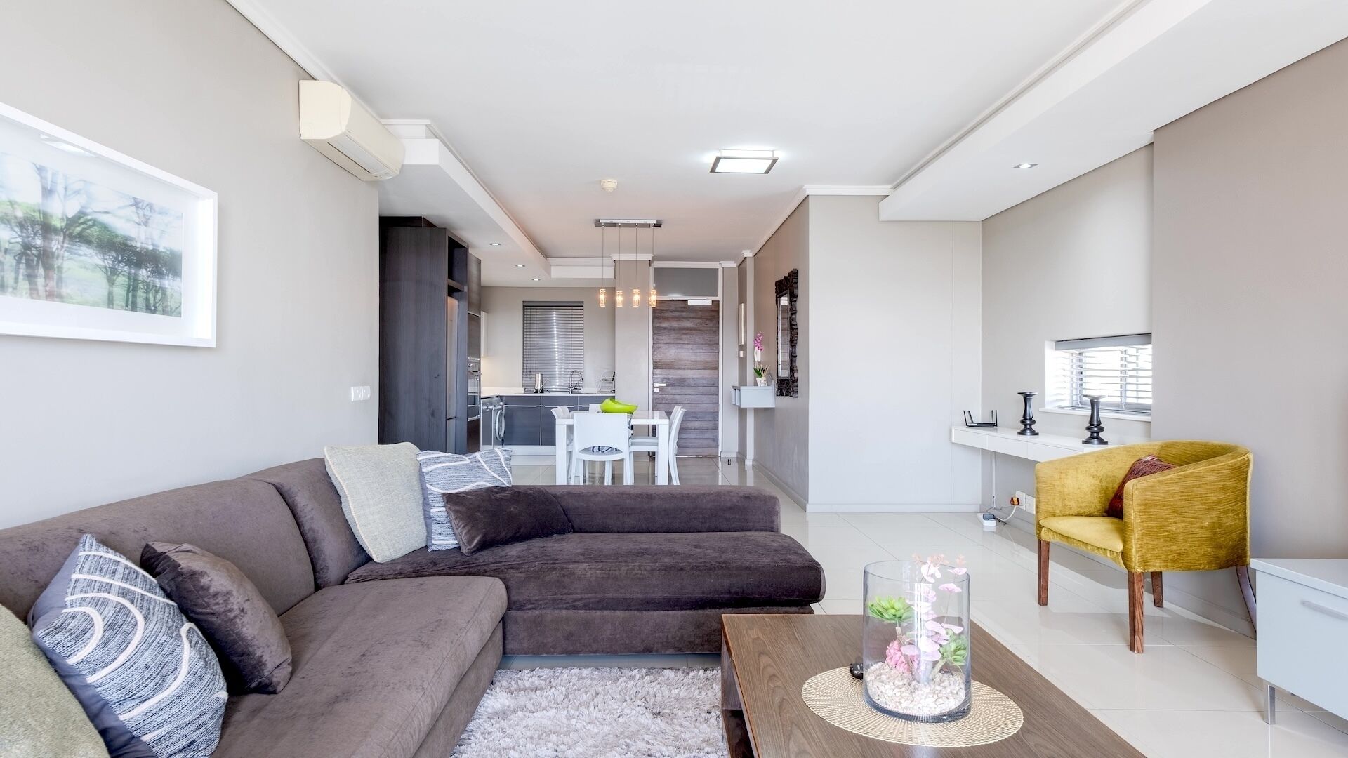 Ein modernes Wohnzimmer mit einer Couch, einem Couchtisch und einem gelben Sessel. Im Hintergrund ist eine Essecke mit einem weißen Tisch und Stühlen zu sehen.
