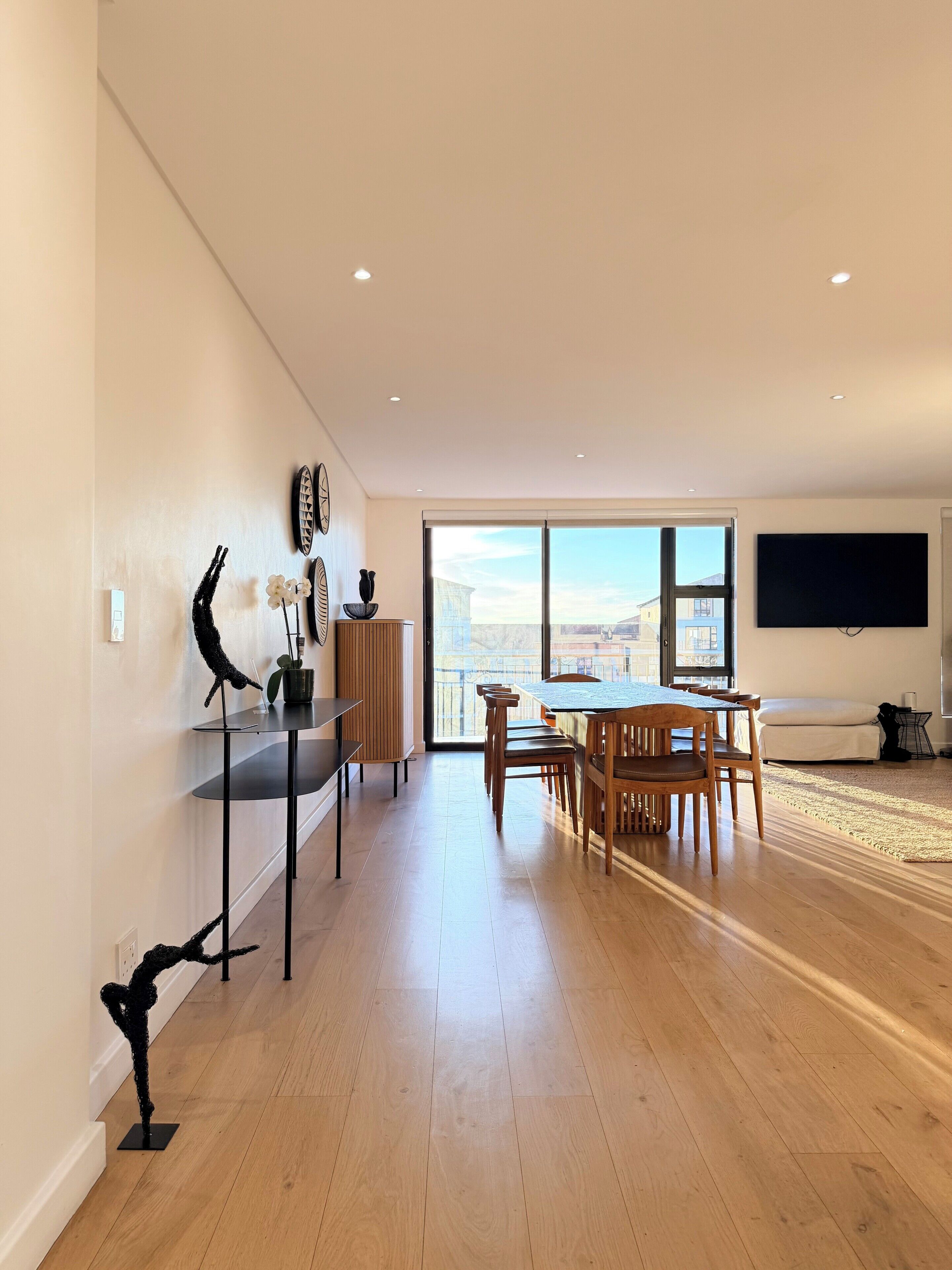 A spacious living area with a dining table, chairs, a sofa, and a large window offering a city view.