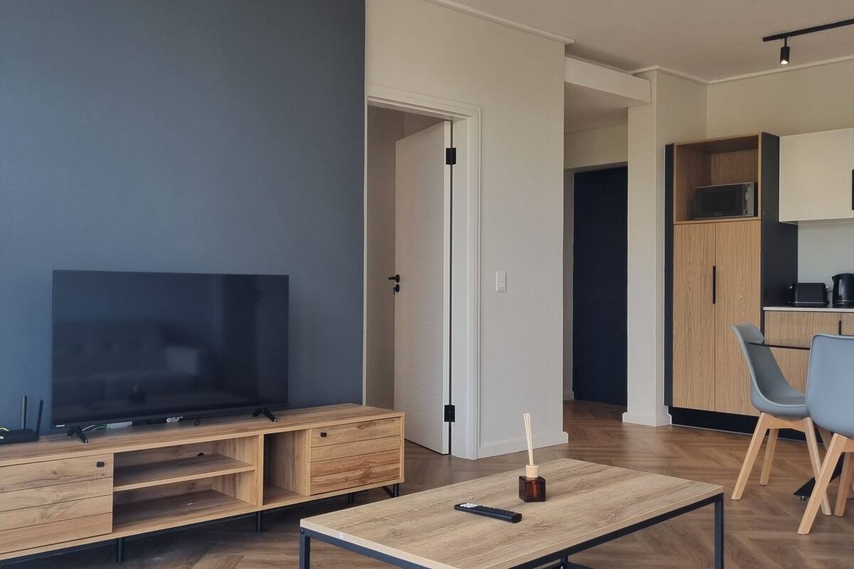 Moderni olohuone, jossa on televisio, puinen mediakeskus, sohvapöytä ja taustalla keittiö.