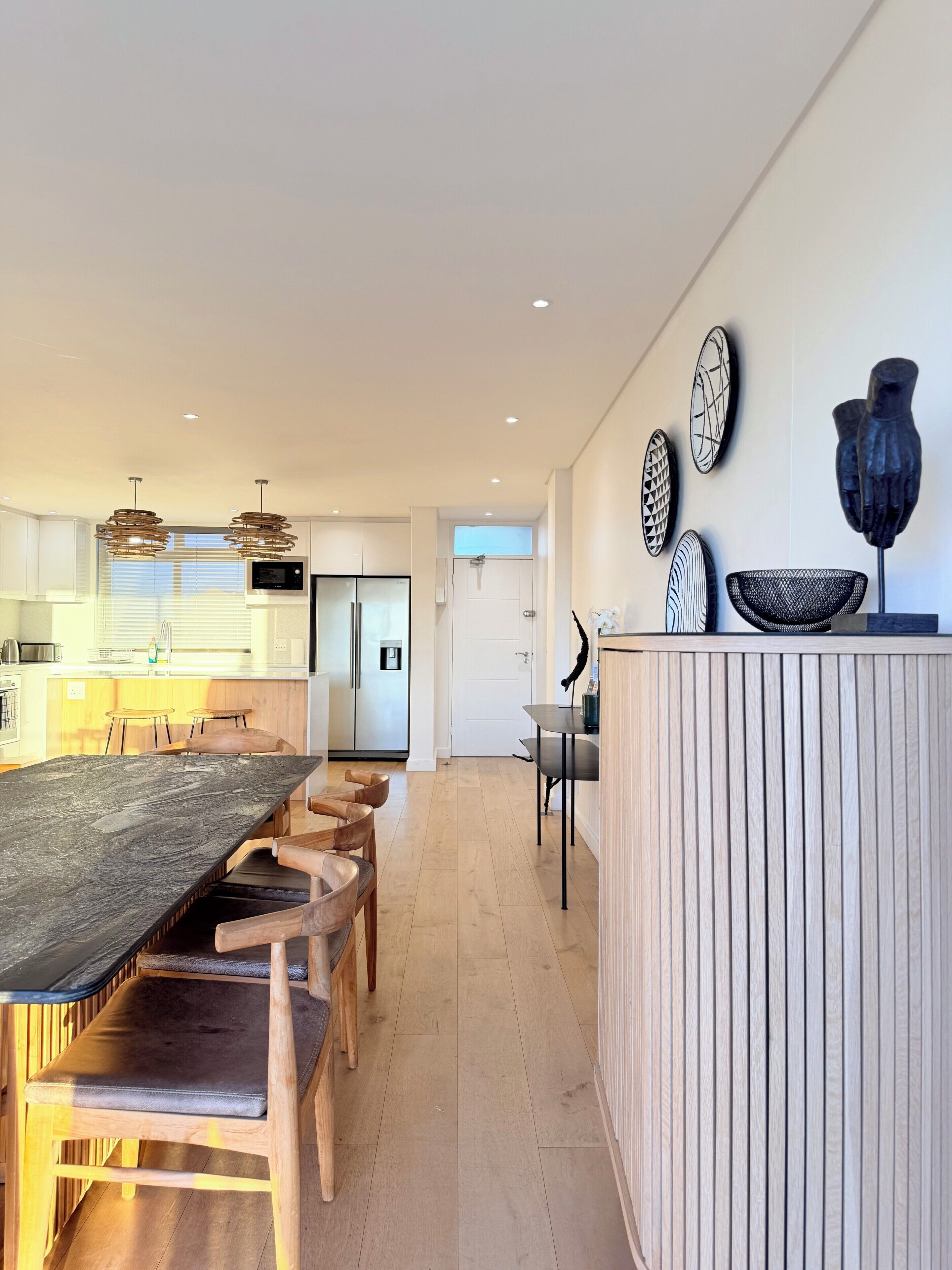 A modern kitchen with a dining area, wooden chairs, a long table, and a white cabinet with decorative items.