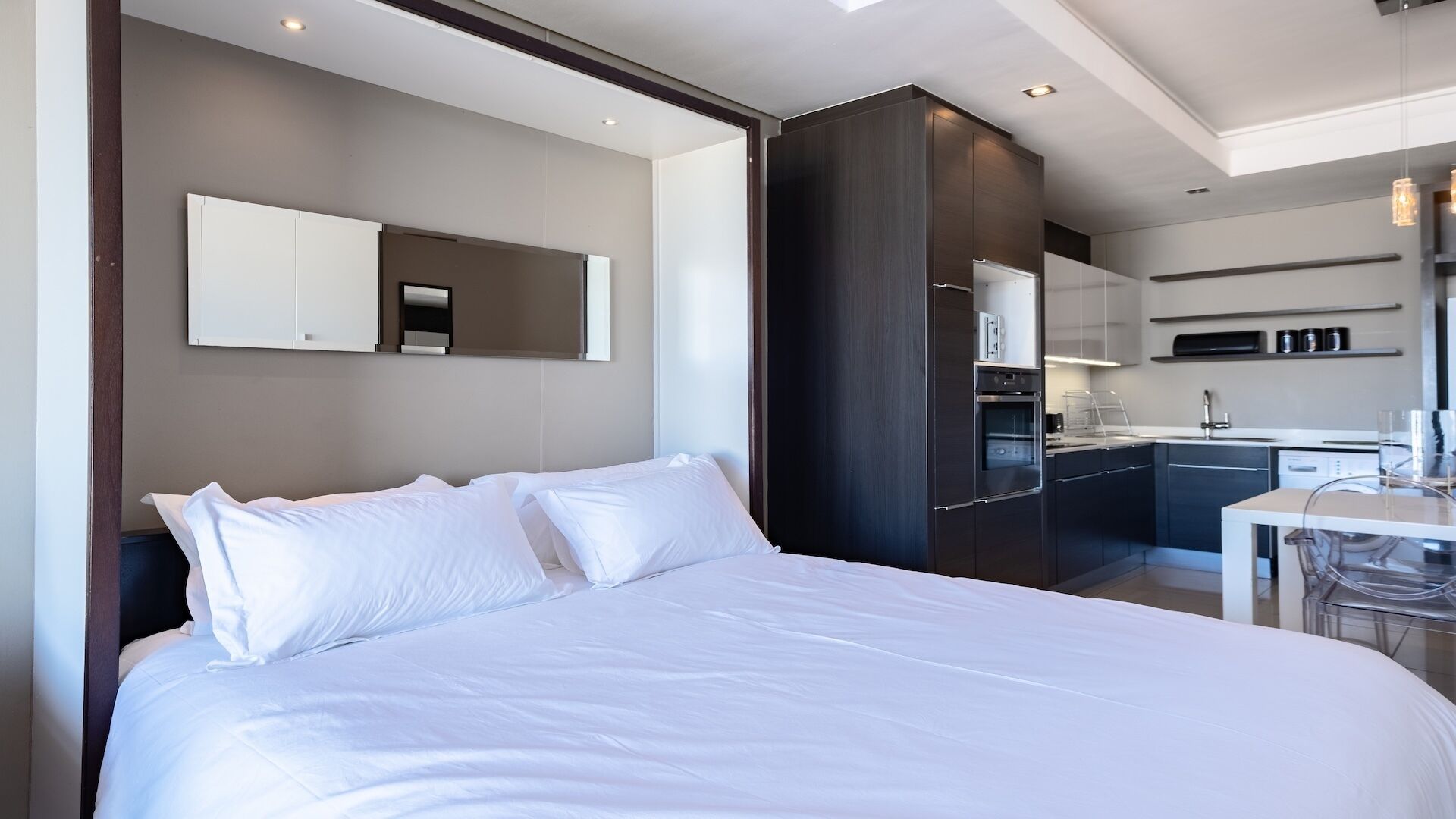 Una habitación de hotel moderna con una cama grande, una cocina integrada con refrigerador y horno, y un pequeño comedor.
