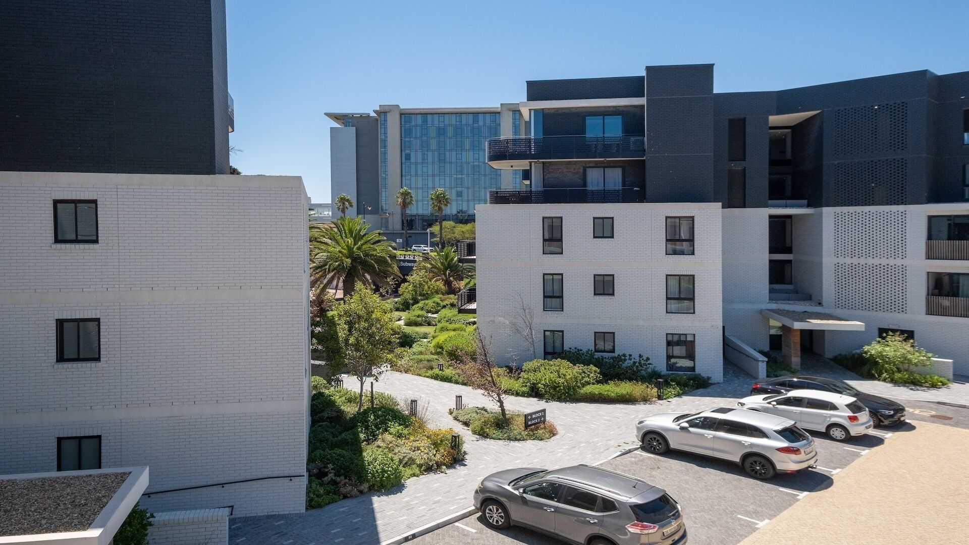 Un immeuble moderne à plusieurs étages, doté d’un parking, où se trouvent plusieurs voitures, et un ciel dégagé.