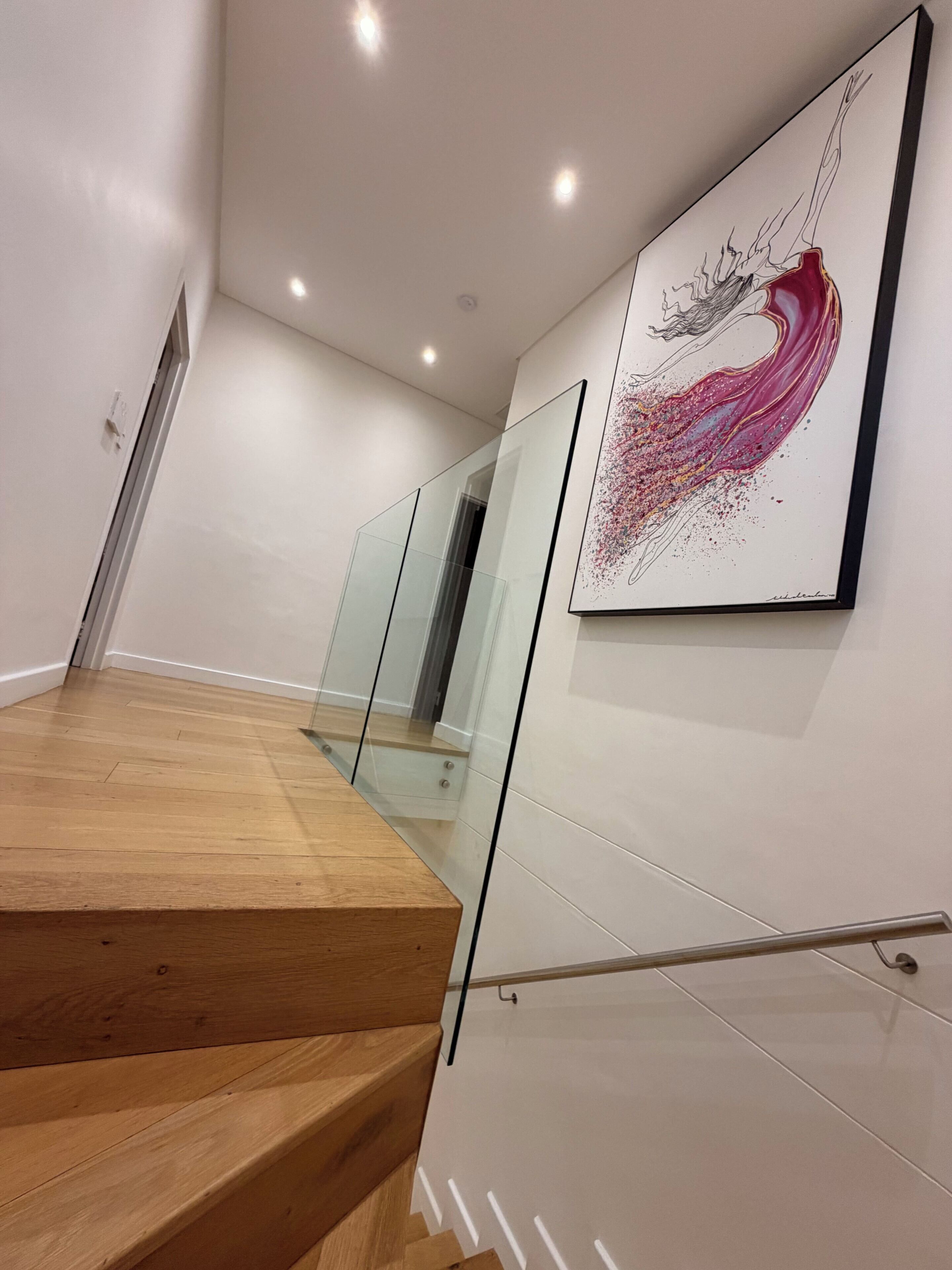 A modern staircase with wooden steps and glass railings, leading to a room with white walls and a large abstract painting.