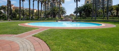 A swimming pool surrounded by a brick walkway, palm trees, and residential buildings in the background.