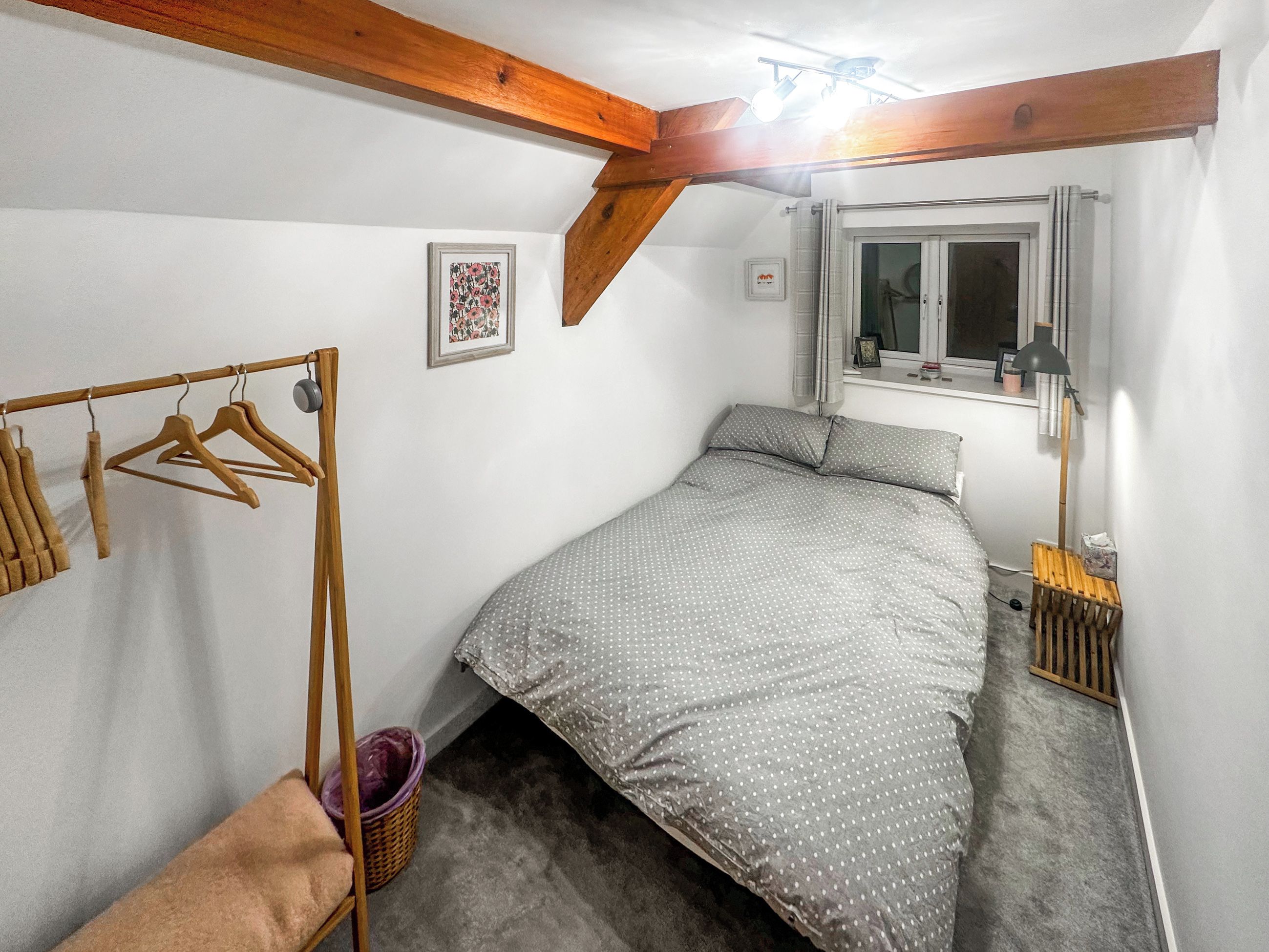 Ein Schlafzimmer mit einem Bett, Holzbalken, einem Fenster, einem Kleiderständer und einem kleinen Korb.