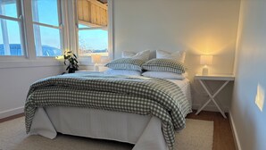 A neatly made bed with a checkered bedspread, a bedside table with a lamp, and a view of the outdoors through a large window. - The Farmhouse Dunedoo (Dunedoo)