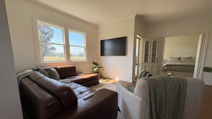 A living room with a brown leather sofa, a white armchair, a flat-screen TV mounted on the wall, and a large window with a view of the outdoors. - The Farmhouse Dunedoo (Dunedoo)