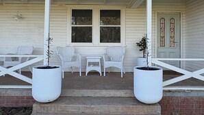 A porch with white wicker chairs and tables, two large potted plants, and a brick pathway leading to the entrance. - The Farmhouse Dunedoo (Dunedoo)