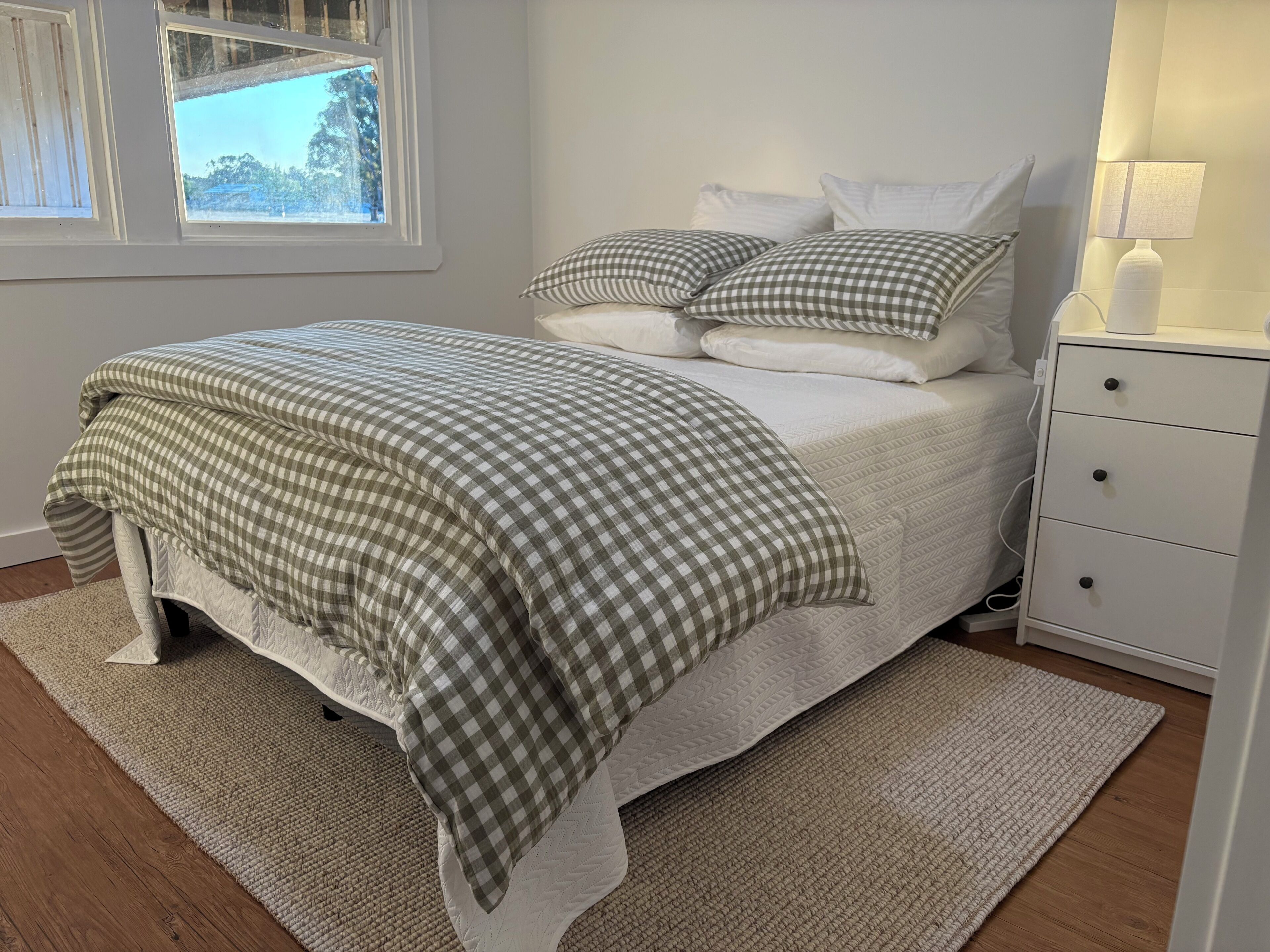 A neatly made bed with a checkered bedspread, a white nightstand with a lamp, and a window with a view of trees.