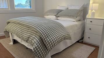 A neatly made bed with a checkered bedspread, a white nightstand with a lamp, and a window with a view of trees.