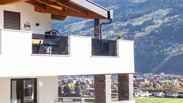 Ein modernes, zweistöckiges Gebäude mit Balkon, Blick auf eine gebirgige Landschaft und einer kleinen Stadt im Hintergrund.