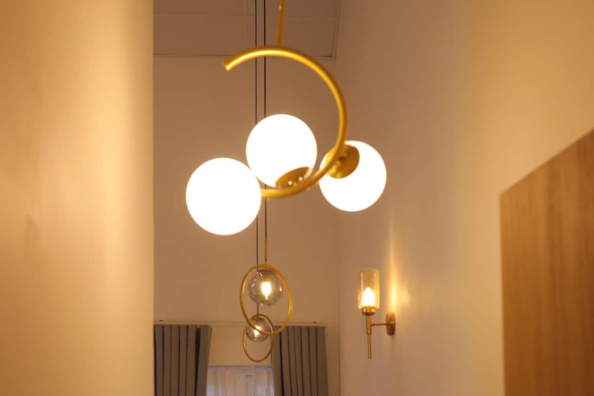 A hallway with a wooden floor, a bedside table with a lamp, a window with curtains, and a wall-mounted light fixture.
