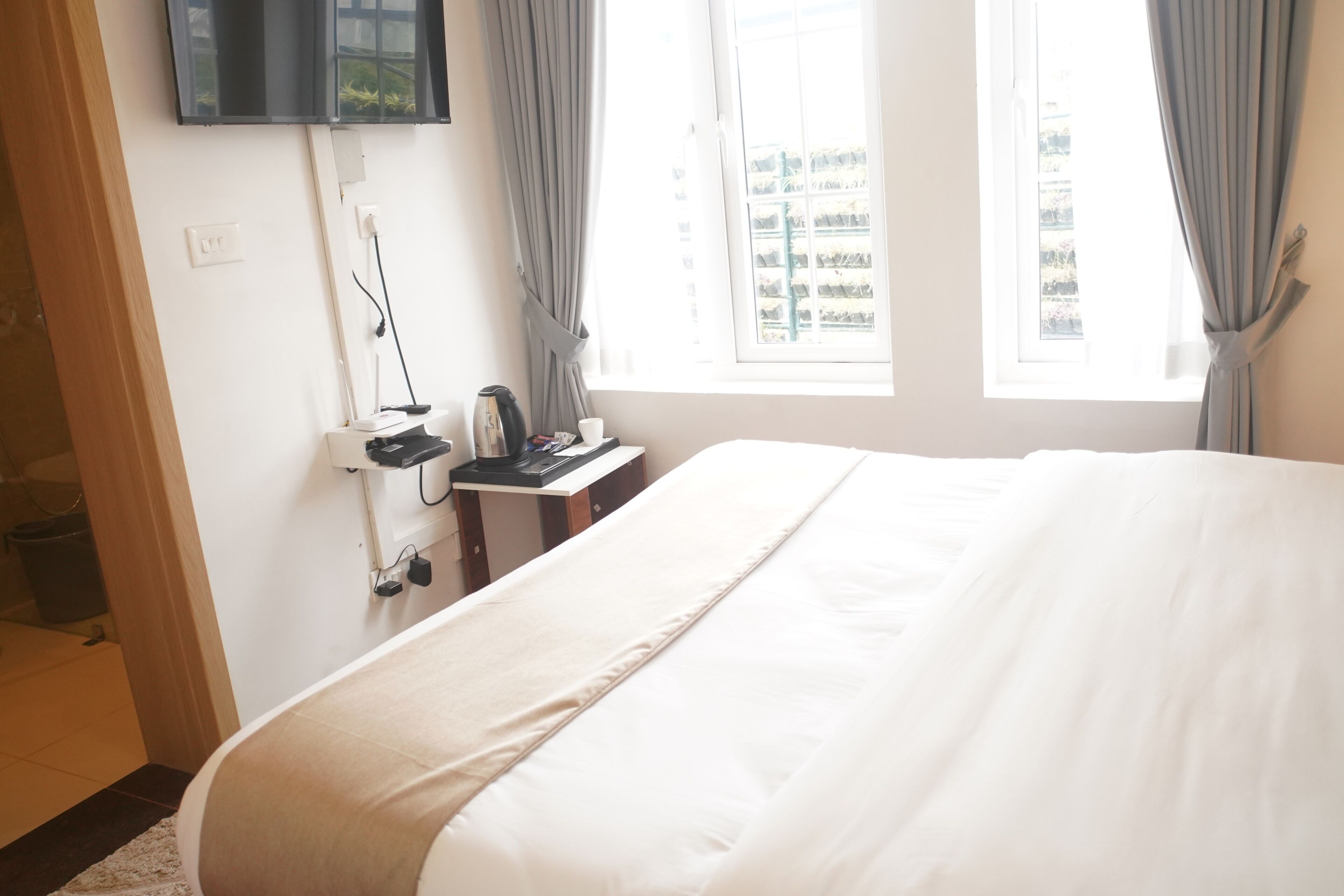 A hotel room with a bed, a flat-screen TV mounted on the wall, a small table with a kettle and a cup, and a window with curtains.