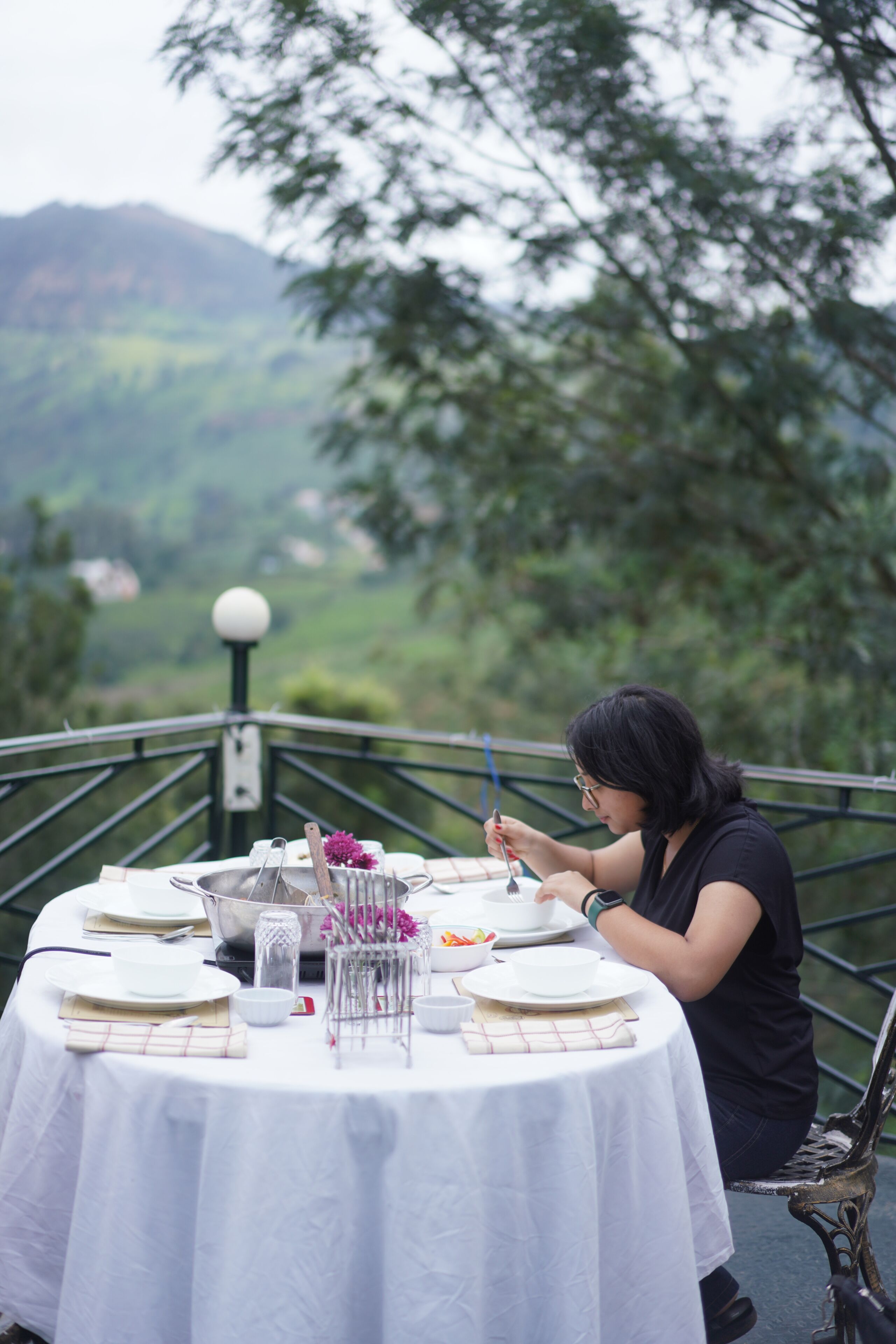 Mesa de comedor al aire libre con una persona sentada, rodeada de platos, cubiertos y un recipiente de vidrio.