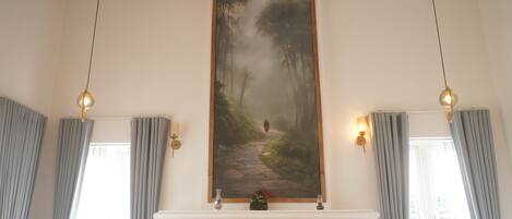 A room with a fireplace, a painting of a forest path, and two wall-mounted lamps.