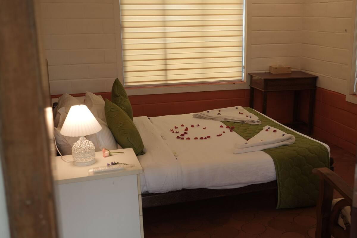 A neatly made bed with a white comforter, green pillows, and a floral arrangement on the bed. There is a bedside table with a lamp and a small vase with flowers.