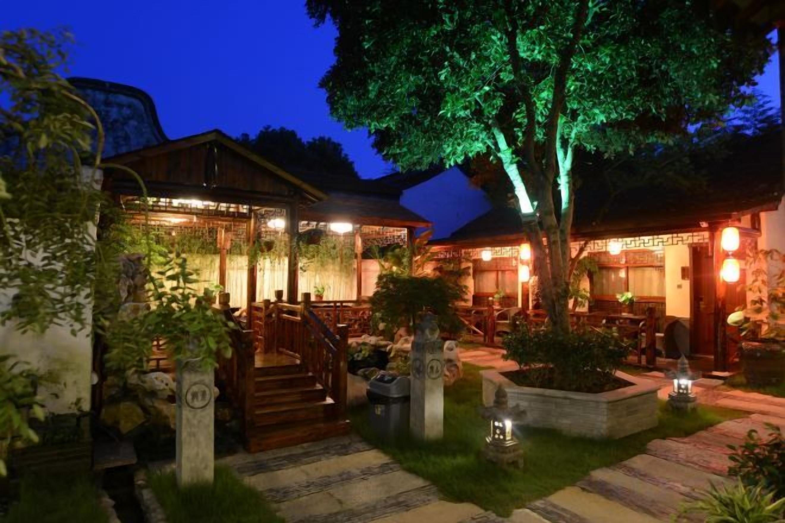 A traditional wooden building with a staircase and outdoor seating area, surrounded by greenery and illuminated by ambient lighting.