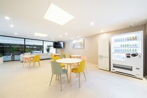 A modern indoor space with round tables and chairs, a vending machine, and a large window with a view of the outdoors. - Hotel Urbanstay (Jeonju)