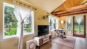 Sala de estar con televisor, cortinas blancas, techo de madera y vista a zonas verdes.