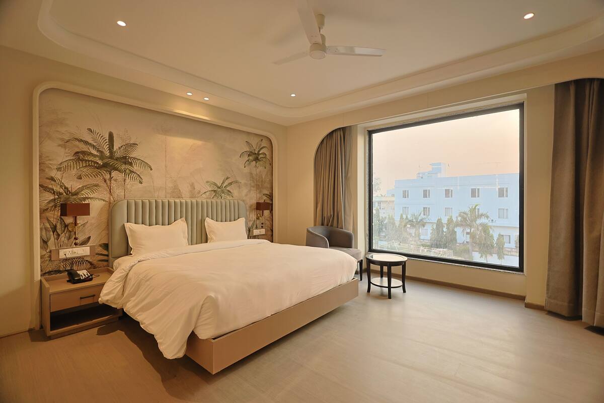 Une chambre d’hôtel moderne avec un grand lit, une petite table ronde, un ventilateur de plafond et une grande fenêtre donnant sur des immeubles et des palmiers.