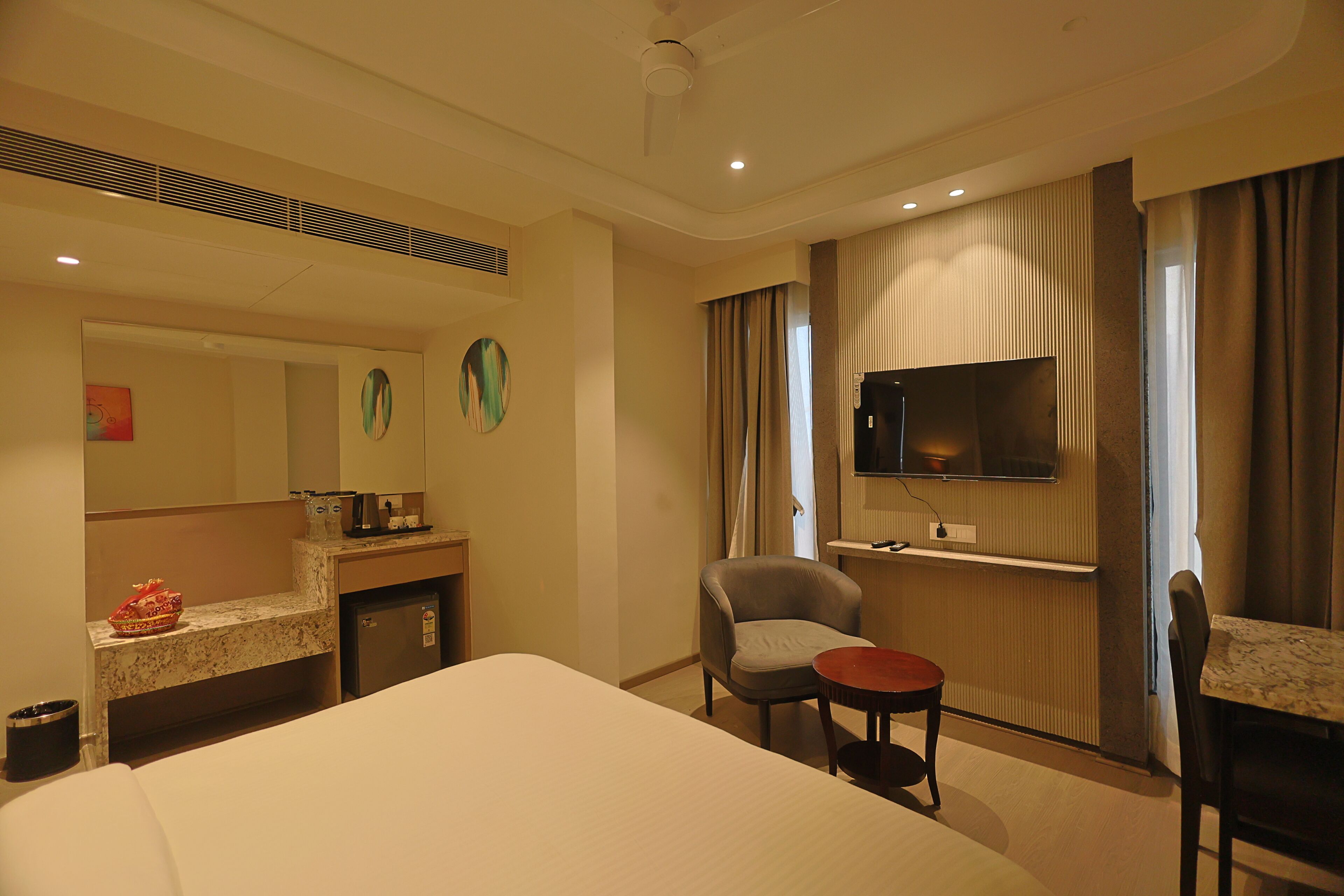 A hotel room with a bed, a small table, a chair, a TV, and a kitchenette area.