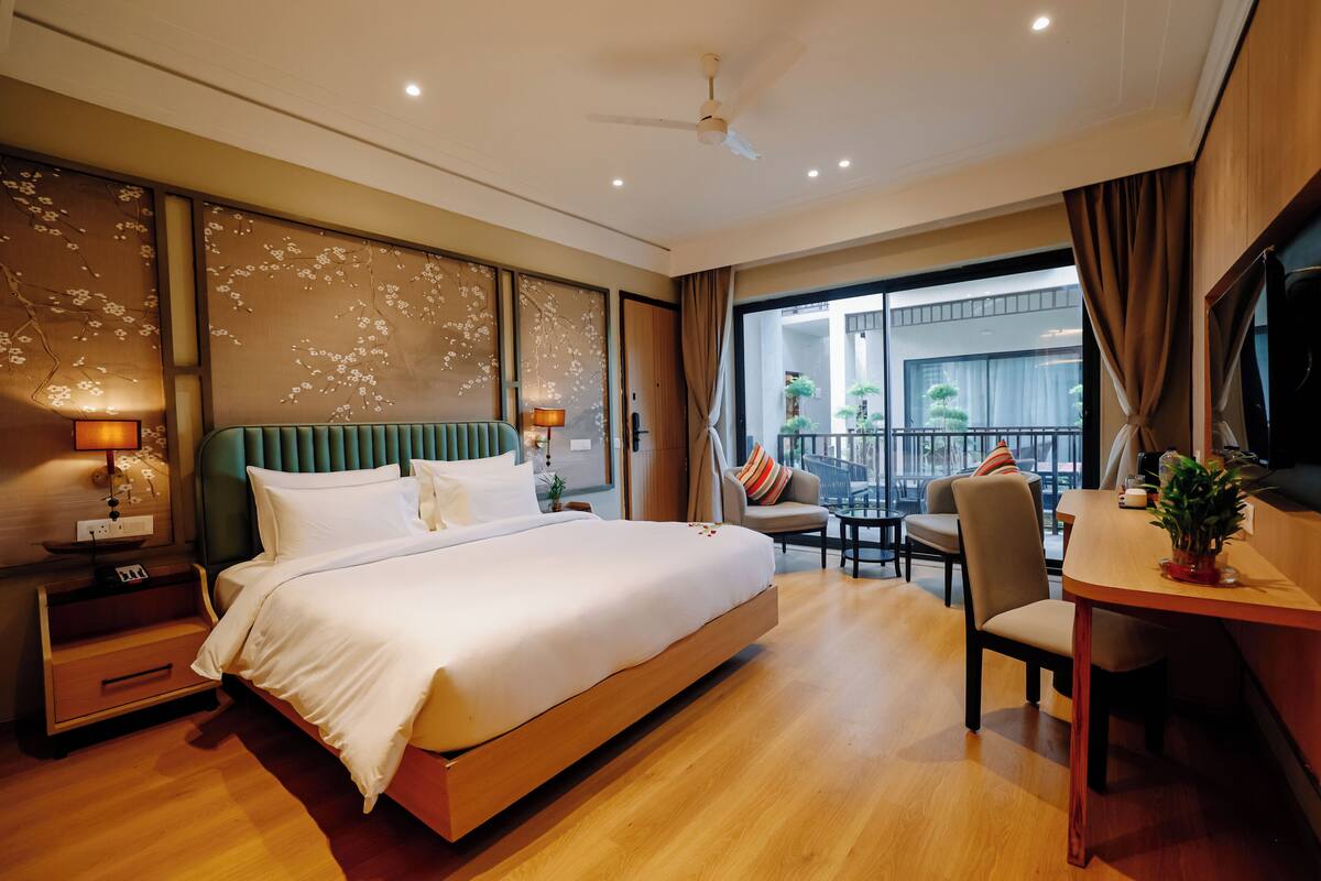 Une chambre d’hôtel avec un grand lit, un bureau, une chaise, une télévision et un balcon agrémenté de plantes.