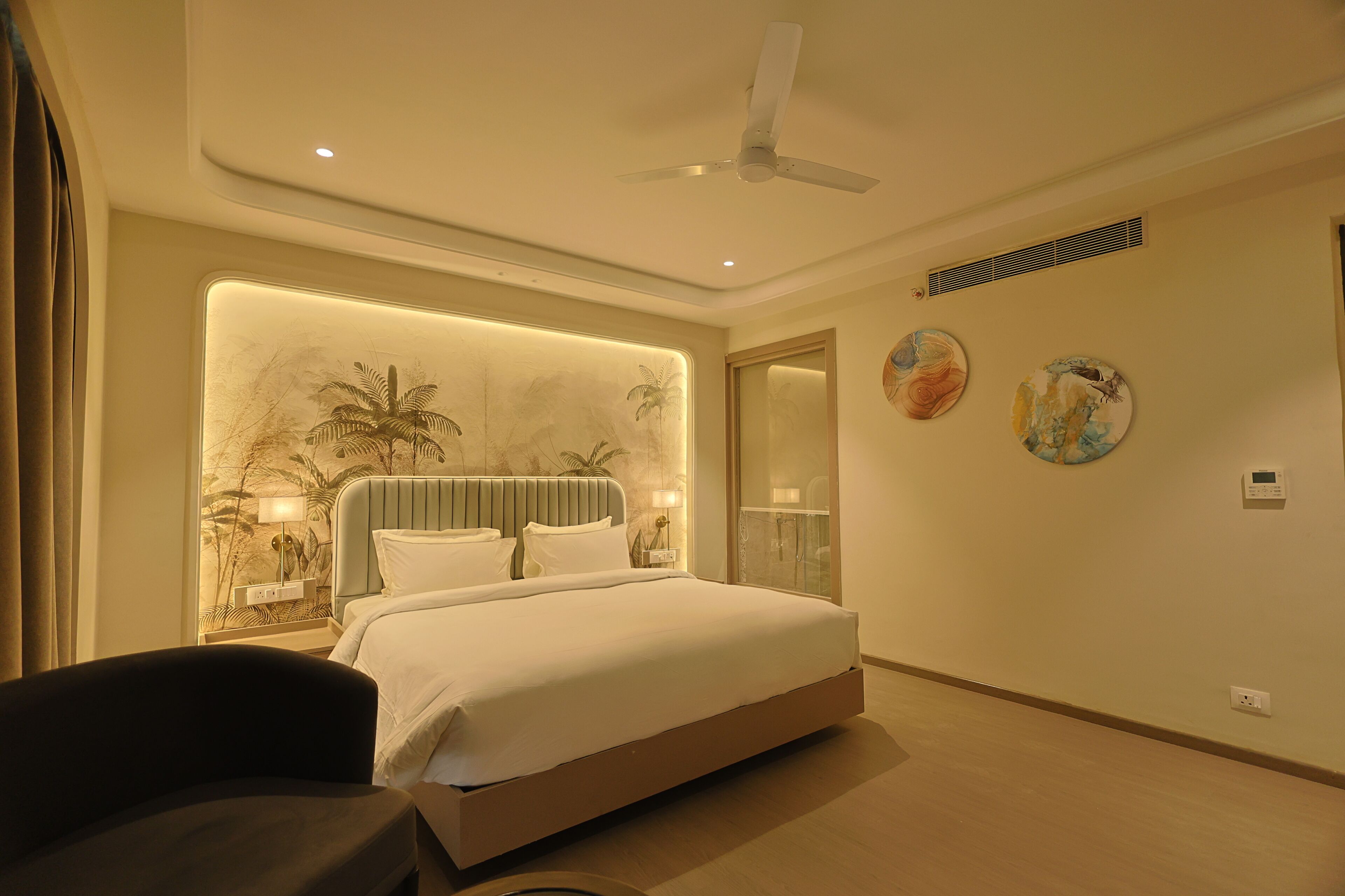 A modern hotel room with a large bed, a ceiling fan, wall art, and a nightstand with a lamp.