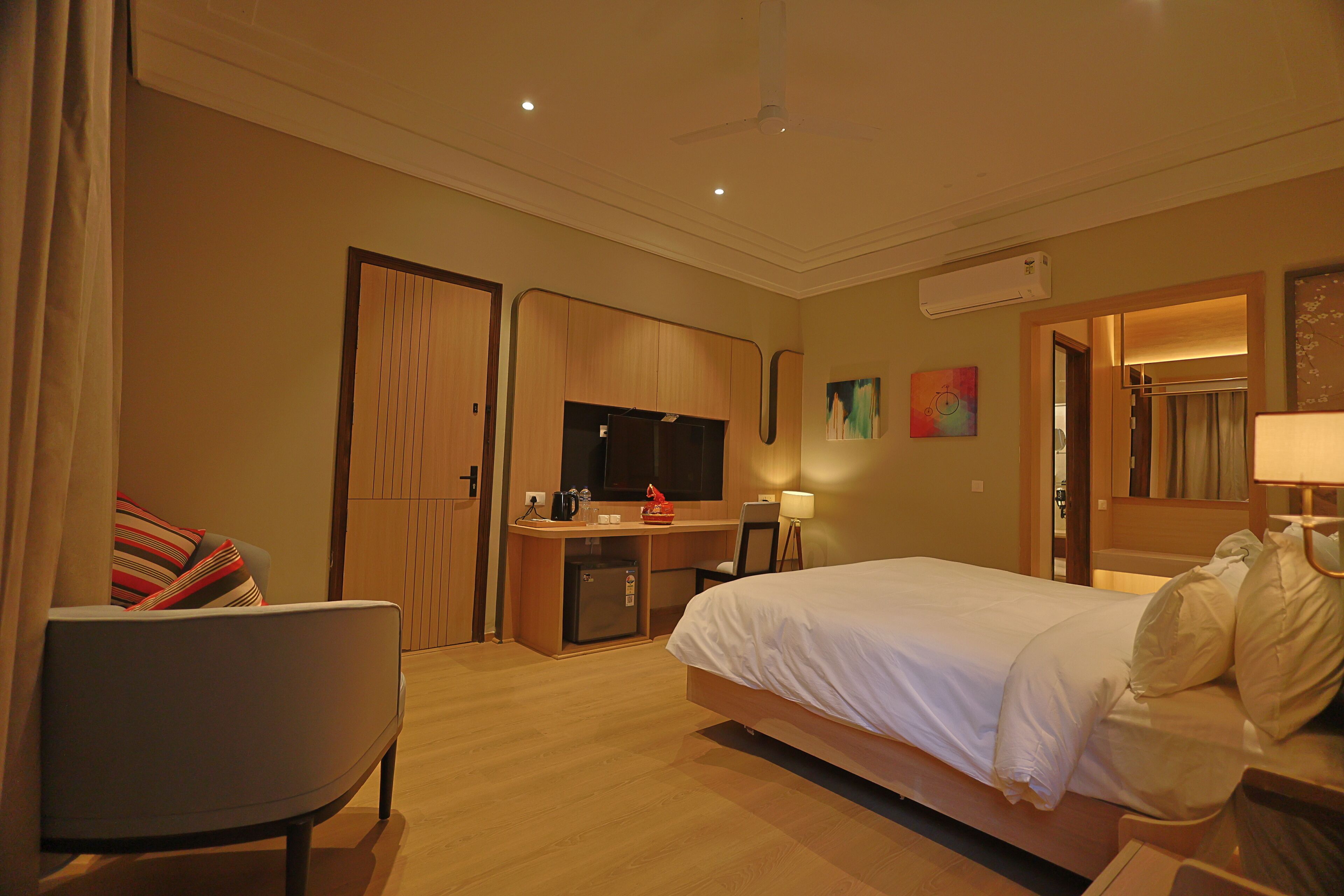 Quarto de hotel com cama, escrivaninha, cadeira, televisão e um pequeno frigorífico.