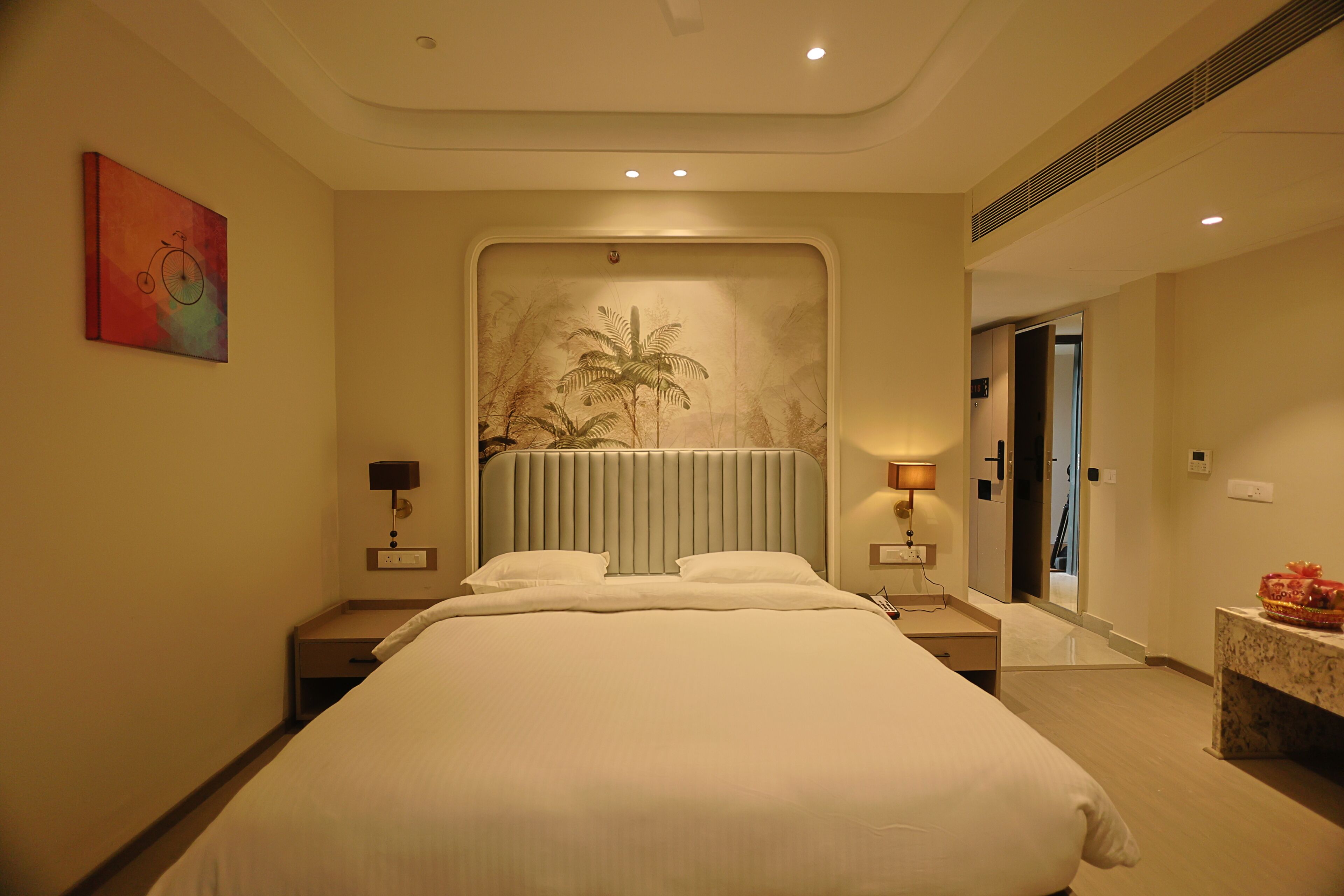 A hotel room with a large bed, two bedside tables with lamps, a wall-mounted artwork, a large mirror, and a door leading to another room.