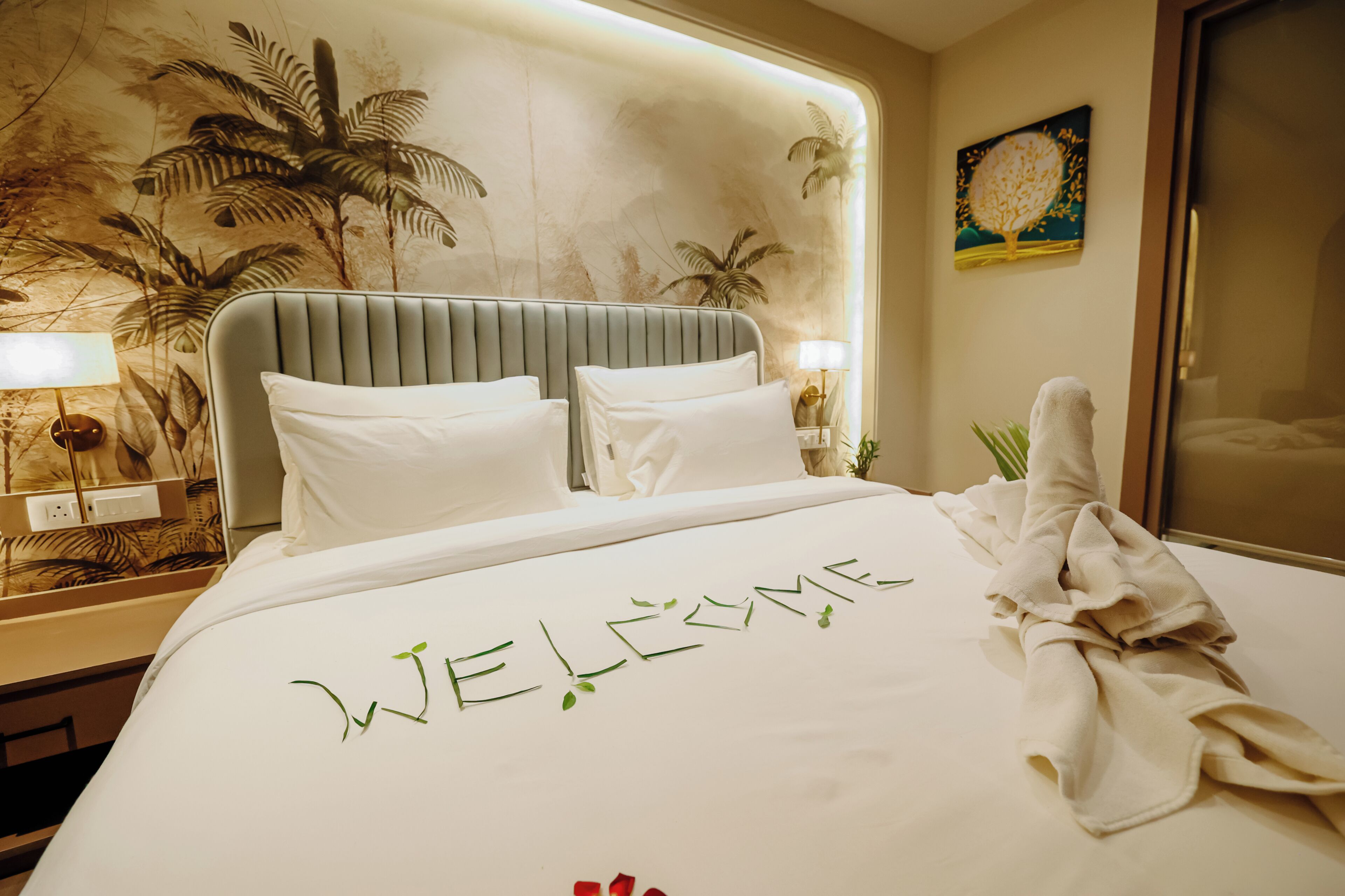 A hotel room with a bed, a headboard, a nightstand, a lamp, a painting, and a towel on the bed.