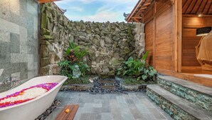 A stone-walled bathroom with a bathtub filled with flowers, a wooden door, and a small garden area with plants. - Aix di Sawah Ubud (Tegallalang)
