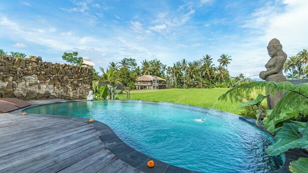 A swimming pool with a wooden deck, a stone wall, and a thatched-roof building surrounded by palm trees. - Aix di Sawah Ubud (Tegallalang)