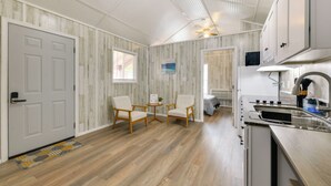 A compact kitchen with a stove, sink, and cabinets. There are two wooden chairs and a small table with a plant. The room has a wooden floor and white walls with a wood grain pattern. - Near Dtwn & Space Center: Tiny Home in Picayune (Picayune)
