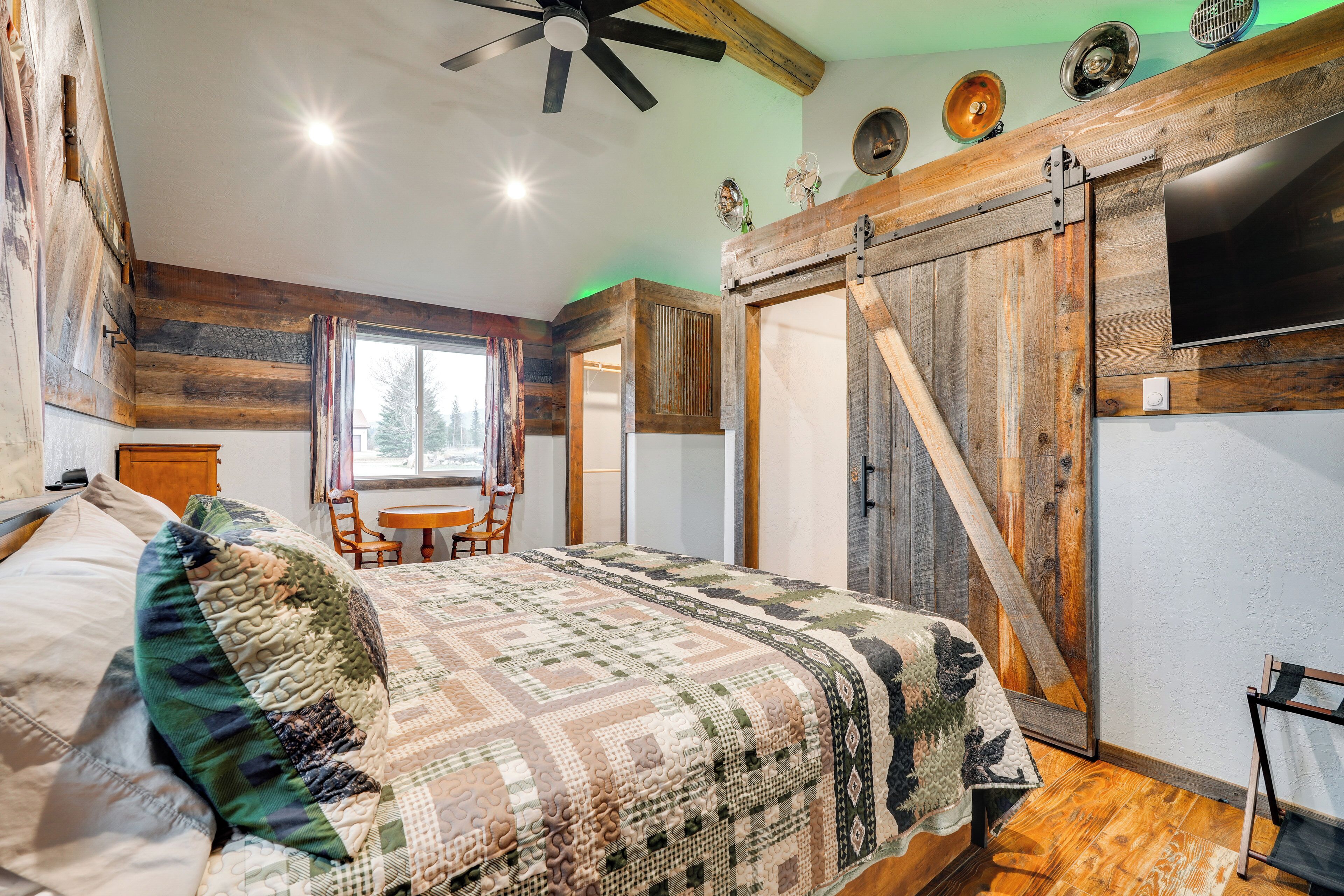 A bedroom with a large bed, a wooden ceiling fan, a sliding barn-style door, and a window with a view of trees.