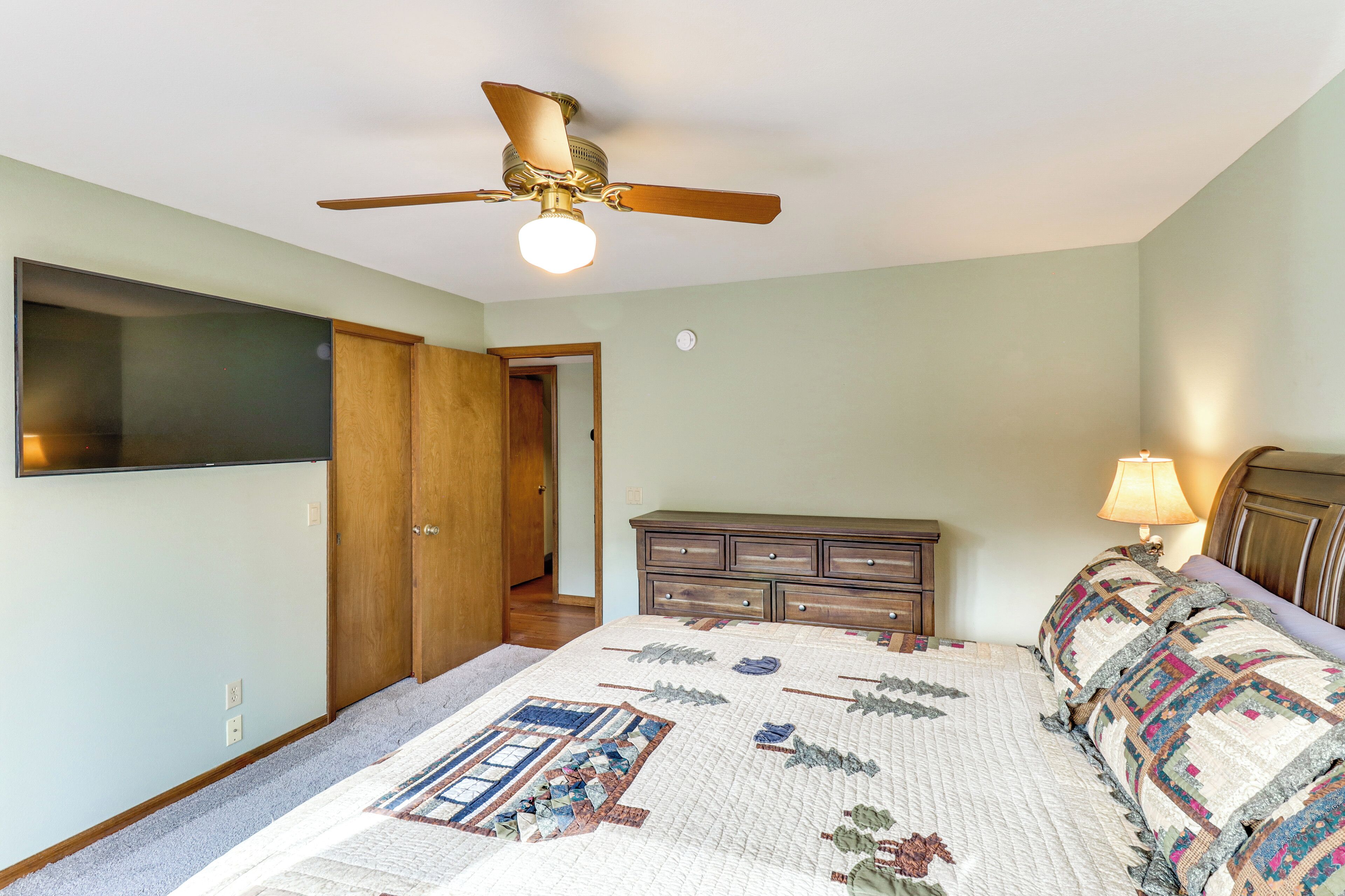 A bedroom with a ceiling fan, a bed with a quilt, a dresser, and a television mounted on the wall.