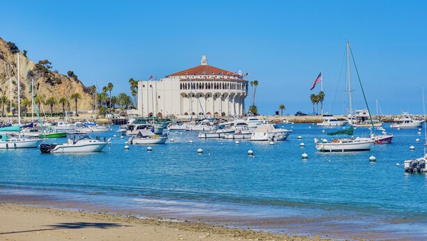 Uma marina com vários barcos atracados, um edifício grande com telhado vermelho e um céu azul claro.