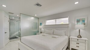 Ein Schlafzimmer mit einem großen Bett, Nachttischen, einem Fenster mit Jalousien und einer Duschkabine aus Glas.