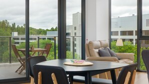 Een moderne woonkamer met een ronde tafel, twee stoelen, een bank en grote ramen met uitzicht op gebouwen en groen.