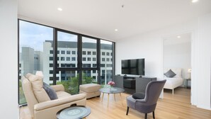 Een moderne woonkamer met een bank, fauteuil en een kleine ronde tafel. Een flatscreen televisie is aan de muur bevestigd, en een bed is op de achtergrond te zien.