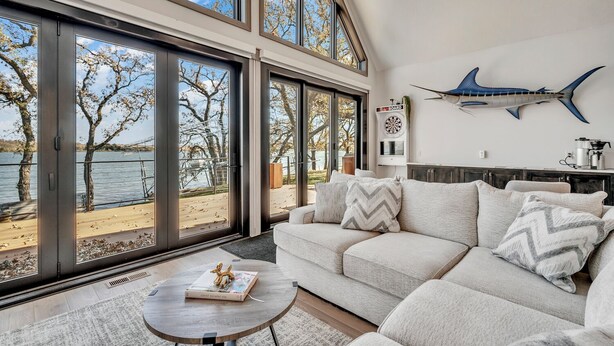 A spacious living room with a large window overlooking a lake, a modern couch, and a dartboard on the wall. - Lower Gar Family Fun- Private Dock, 6 bedrooms, Close to boat Ramp (Arnolds Park)
