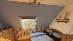 Un dormitorio pequeño y acogedor con techo inclinado, tragaluz, cama con almohadas, cómoda de madera y estante con un modelo de velero.