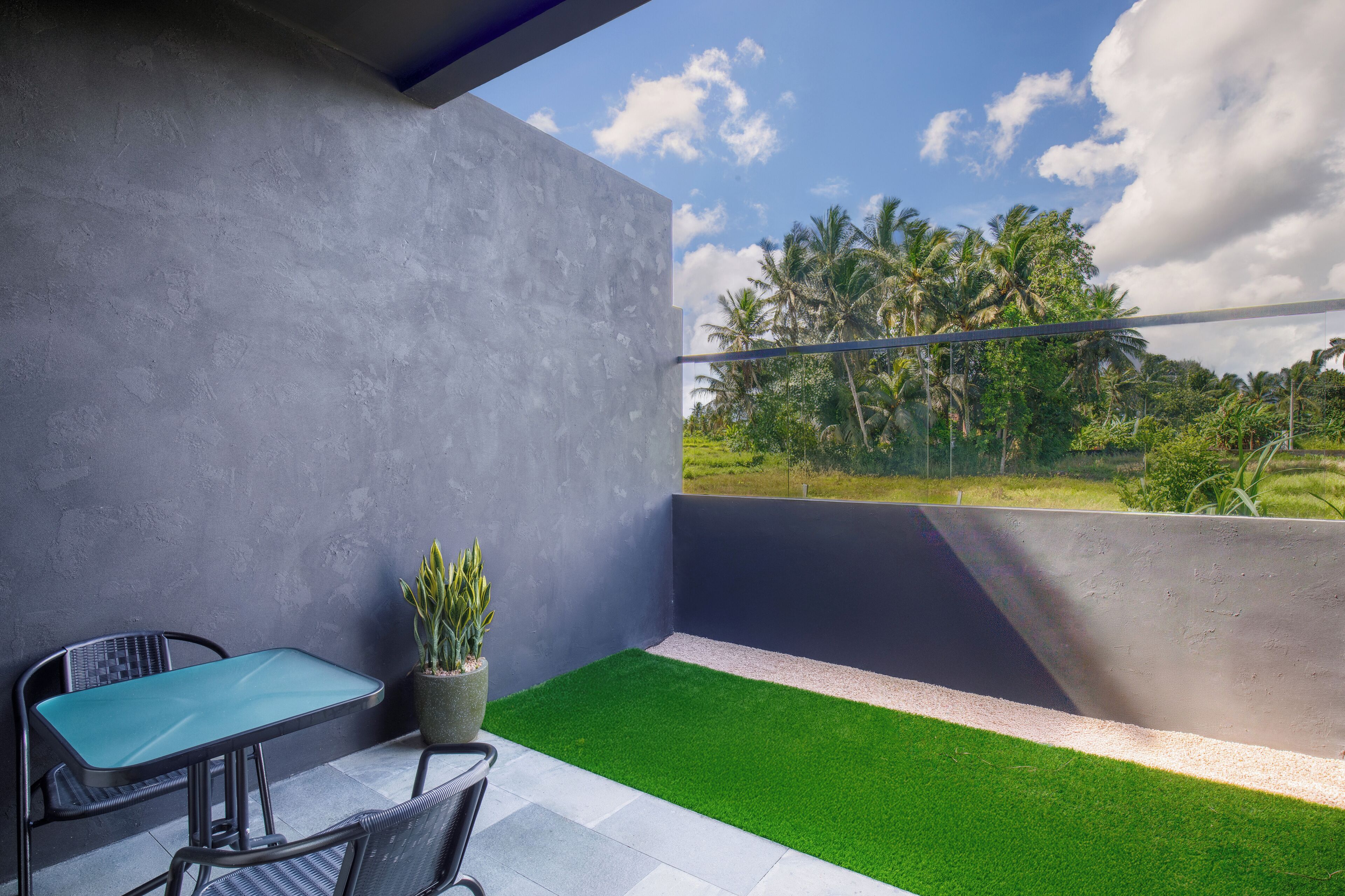 A balcony with a green artificial grass floor, a table, and chairs, overlooking a landscape with palm trees and a clear sky.